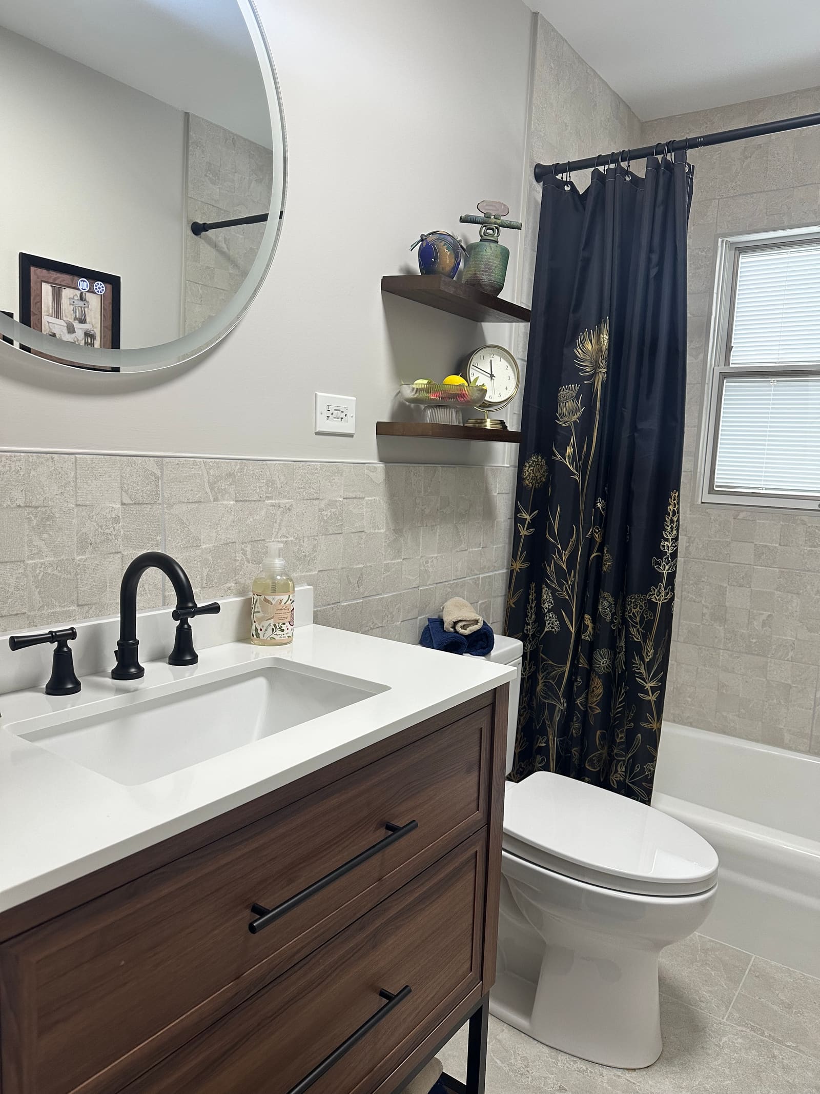 Bathroom with a walnut vanity and white quartz top in the foreground, tiled tub and shower beyond, and a round mirror on the left.